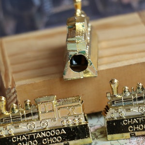 Op de afbeelding: Drie gouden metalen potloodpunten in de vorm van treinen. De potloodpunten zijn gedetailleerd met zwarte lijnen en wielen. De voorkant van elke potloodpunt heeft de tekst "Chattanooga Choo Choo".