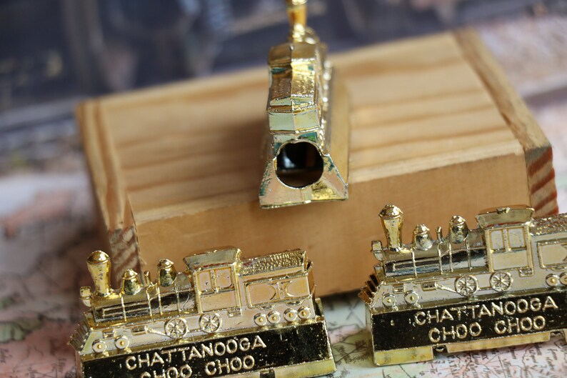Op de afbeelding: Drie gouden metalen potloodpunten in de vorm van treinen. De potloodpunten zijn gedetailleerd met zwarte lijnen en wielen. De voorkant van elke potloodpunt heeft de tekst "Chattanooga Choo Choo".