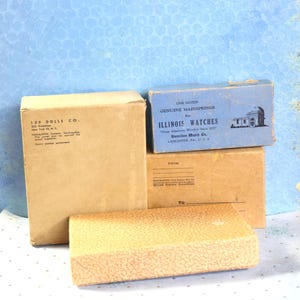 May include: Three cardboard boxes stacked on top of each other. The top box is blue and says "One Dozen Genuine Mainsprings for Illinois Watches "Great American Watches Since 1870" Hamilton Watch Co. Lancaster, PA. U.S.A."