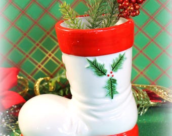 May include: White ceramic Santa boot with red trim and holly design, used as a vase with green pine sprigs and red berries.