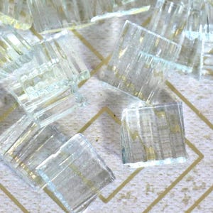 May include: A collection of small, clear, square glass tiles with a textured surface. The tiles are scattered on a white surface with a gold geometric pattern. The glass has a slight blue tint.