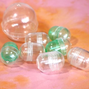 May include: A collection of clear plastic capsule toys, some with green accents, and a larger clear plastic dome. The capsules are scattered on a pink and orange surface. The toys are designed to hold small items.