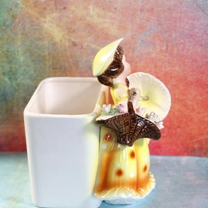 May include: A ceramic planter shaped like a girl holding a turkey. The girl is wearing a yellow dress with brown polka dots and a yellow hat. The planter is white with a yellow and brown design.