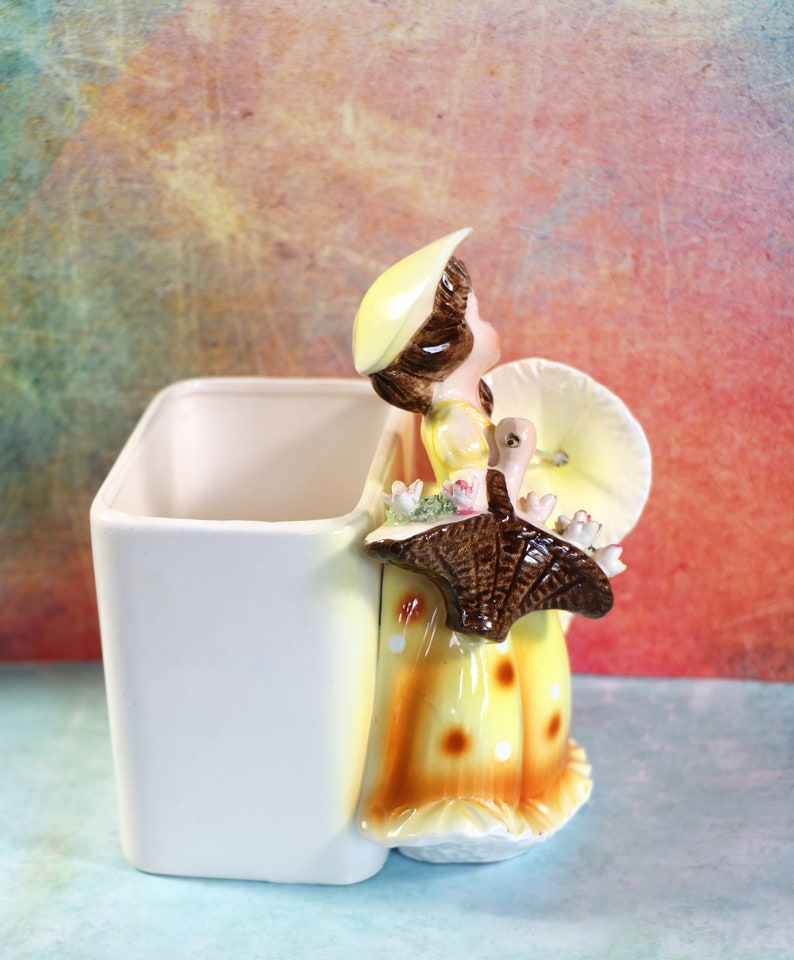 May include: A ceramic planter shaped like a girl holding a turkey. The girl is wearing a yellow dress with brown polka dots and a yellow hat. The planter is white with a yellow and brown design.