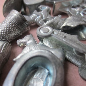 May include: A close-up of a collection of vintage metal game pieces, including a thimble, a car, a horse, and a shoe. The pieces are all silver in color and have a worn, antique look.