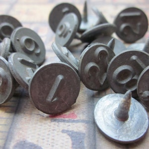 May include: A collection of metal number stamps with a nail on the back. The numbers are 1, 2, 3, 4, 5, 6, 7, 8, and 9. The stamps are silver in color and have a worn, vintage look.