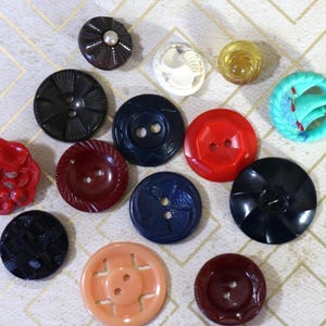 May include: A collection of thirteen assorted sewing buttons in various shapes, sizes, and colors including red, black, blue, peach, and white. The buttons feature diverse textures, patterns, and hole configurations, ranging from floral designs to geometric shapes.