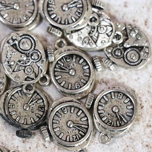 May include: A collection of small, silver-toned clock charms. The charms feature intricate clock face designs with visible hands and numbers, and some show the inner workings of a clock. Each charm has a small loop for attaching to jewelry or other crafts.