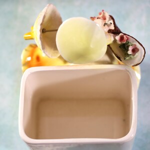 May include: A white ceramic toothbrush holder with a decorative floral design. The holder features a yellow and brown floral design with pink flowers. The holder is empty.