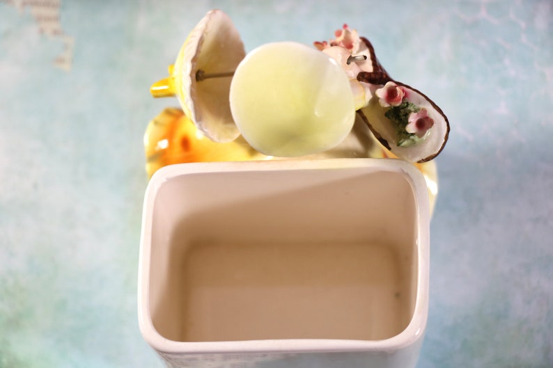 May include: A white ceramic toothbrush holder with a decorative floral design. The holder features a yellow and brown floral design with pink flowers. The holder is empty.