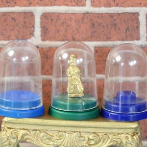 May include: Three clear plastic display domes with colored bases. One dome contains a small gold figurine. The bases are blue and green. The domes sit on a decorative gold-colored stand against a brick wall.