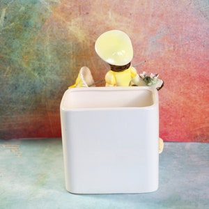 May include: White ceramic planter with a figurine of a girl wearing a yellow dress and a yellow hat. The girl is holding a paintbrush and a palette.