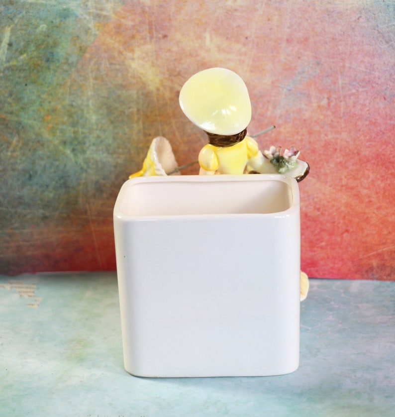 May include: White ceramic planter with a figurine of a girl wearing a yellow dress and a yellow hat. The girl is holding a paintbrush and a palette.