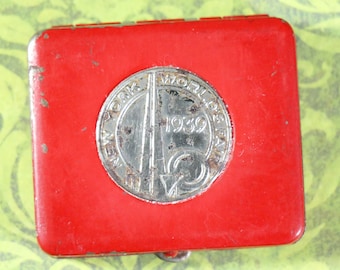 May include: Red metal compact with a silver circular emblem featuring the text "New York World's Fair 1939".