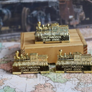 Op de afbeelding: Drie gouden miniatuur stoomlocomotieven met de woorden "Chattanooga Choo Choo" op de onderkant van elk. De locomotieven staan op een houten oppervlak en een wereldkaart.