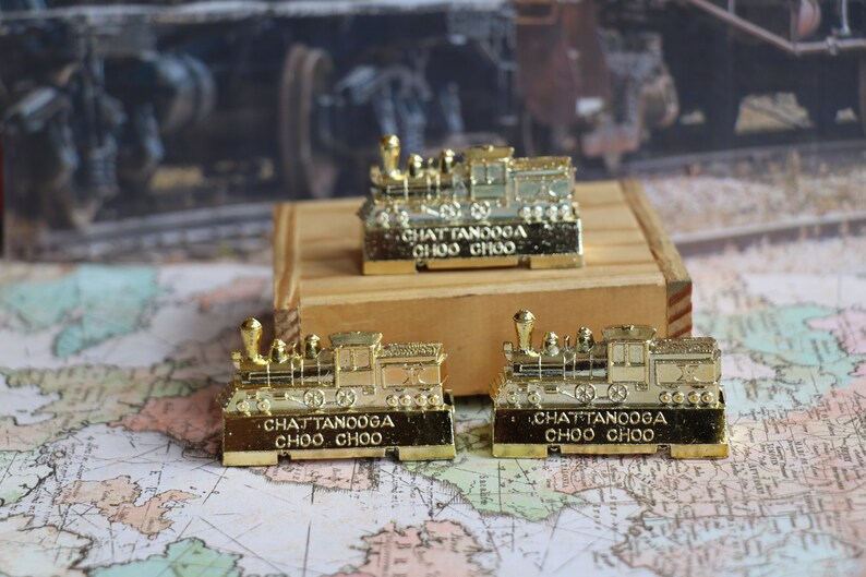 Op de afbeelding: Drie gouden miniatuur stoomlocomotieven met de woorden "Chattanooga Choo Choo" op de onderkant van elk. De locomotieven staan op een houten oppervlak en een wereldkaart.