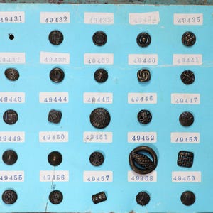 May include: A collection of antique bronze buttons arranged on a light blue display board. Each button has a unique design, including geometric patterns and floral motifs. The board has labels with numbers.