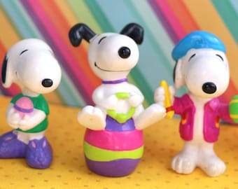 May include: Three small plastic figurines of Snoopy, each in a different pose and holding a different Easter-themed item. One Snoopy is holding a purple and white egg, another is holding a yellow and green egg, and the third is holding a yellow and green pencil.