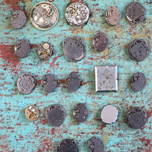 May include: A collection of 20 watch parts and mechanisms arranged on a blue and green textured surface. The watch parts are silver, gold, and black in colour.