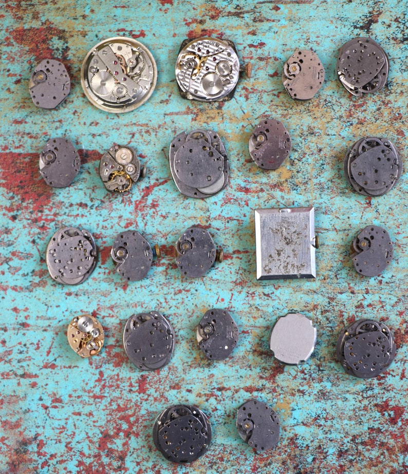 May include: A collection of 20 watch parts and mechanisms arranged on a blue and green textured surface. The watch parts are silver, gold, and black in colour.
