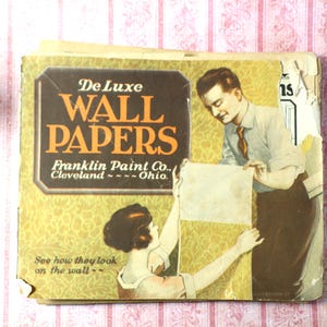 May include: Vintage "De Luxe Wall Papers" catalog from Franklin Paint Co. in Cleveland, Ohio. The cover features an illustration of a man and woman applying wallpaper, with the text "See how they look on the wall." The catalog is aged with visible wear.