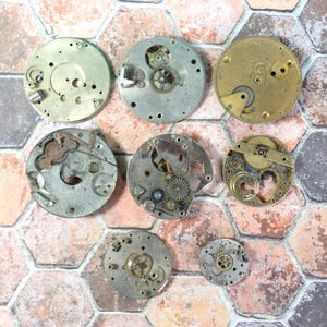 May include: A collection of vintage watch movement parts, including gears, plates, and other components. The parts are in various shapes and sizes, with a mix of silver and gold tones. The arrangement is on a hexagonal brick background.