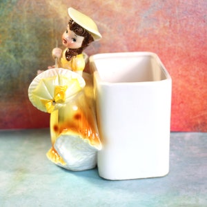 May include: A ceramic figurine of a girl in a yellow dress and hat holding an umbrella. The figurine is attached to a white rectangular planter.