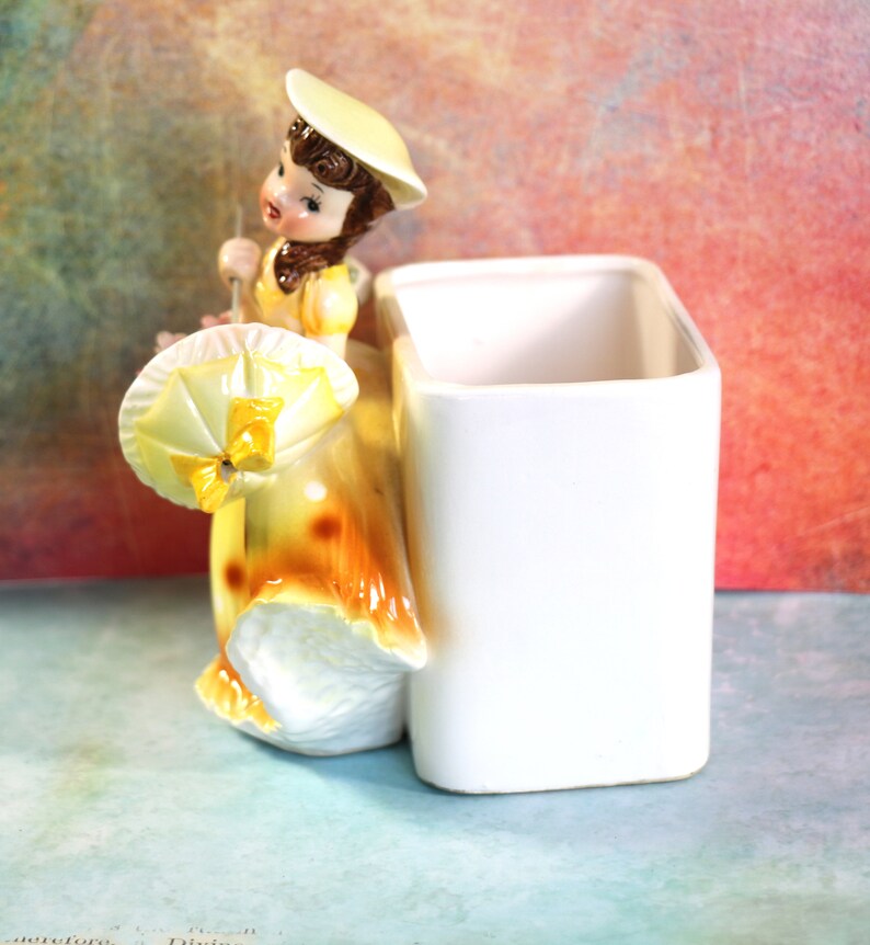 May include: A ceramic figurine of a girl in a yellow dress and hat holding an umbrella. The figurine is attached to a white rectangular planter.