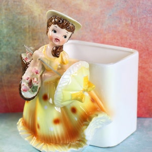 May include: A vintage ceramic planter shaped like a girl in a yellow dress with white polka dots, holding a yellow umbrella and a basket of pink flowers. The planter has a white square base.