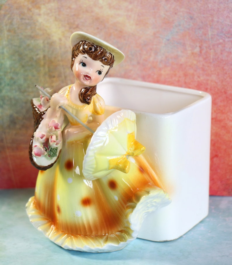 May include: A vintage ceramic planter shaped like a girl in a yellow dress with white polka dots, holding a yellow umbrella and a basket of pink flowers. The planter has a white square base.