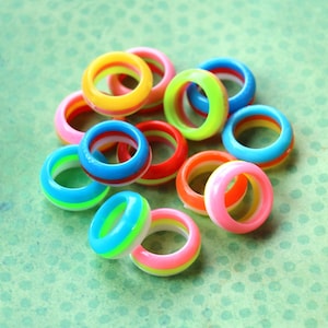 May include: A collection of colorful resin rings in various shades of pink, orange, yellow, green, and blue. The rings are stacked together, creating a vibrant display of accessories. The rings are approximately 1 inch in diameter.