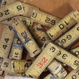 May include: A collection of vintage yellow folding ruler segments scattered in a pile. Each wooden segment features black printed imperial measurements in inches, with some numbers highlighted in red ink, showing various numerical increments.