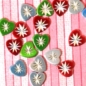 May include: A collection of heart-shaped embellishments in various colors, including red, green, and blue-gray. Each heart features a starburst design in a contrasting color. The embellishments are scattered on a pink and white striped background.