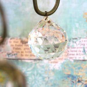 May include: A clear, faceted crystal ball ornament hangs from a dark green metal hook. The crystal reflects light, creating a sparkling effect. The background features a soft, textured pattern with hints of blue and cream.