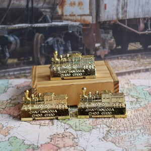 Op de afbeelding: Drie gouden miniatuurtreinlocomotieven met de tekst "Chattanooga Choo Choo" erop gedrukt. De locomotieven staan op een houten oppervlak.