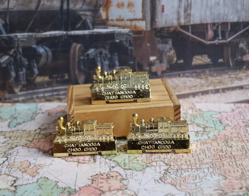 Op de afbeelding: Drie gouden miniatuurtreinlocomotieven met de tekst "Chattanooga Choo Choo" erop gedrukt. De locomotieven staan op een houten oppervlak.