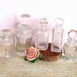 May include: A collection of vintage glass perfume bottles with decorative stoppers. The bottles vary in shape and size, with clear glass and a few with etched details. A ceramic rose and a wooden base add to the display.
