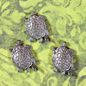 May include: Three silver turtle charms with detailed shell designs. The turtles are facing upwards with their legs and heads visible.
