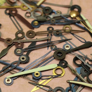 May include: A collection of assorted watch hands in various colors and styles, including gold, silver, black, and white. The hands are made of metal and have different lengths and shapes.