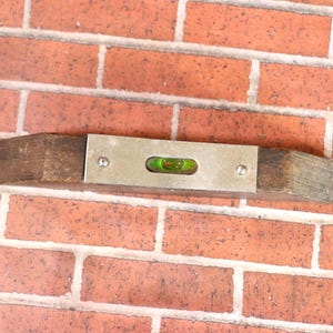 May include: A vintage wooden level with a metal plate and a green bubble vial. The level is mounted on a red brick wall, showcasing its use in construction or carpentry. The tool is designed to ensure surfaces are perfectly horizontal.