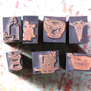 May include: Eight vintage wooden printing blocks with raised designs. The designs include a mortar and pestle, a bottle, a snake, a stylized letter, and the word "Pharmacy".