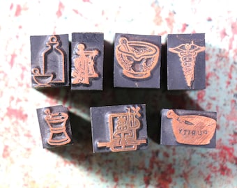 May include: Eight vintage wooden printing blocks with raised designs. The designs include a mortar and pestle, a bottle, a snake, a stylized letter, and the word "Pharmacy".