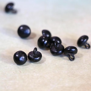 May include: A collection of small, round, black buttons with a glossy finish. Each button has a loop on the back for sewing. The buttons are scattered on a light-colored surface, with some in focus and others blurred.