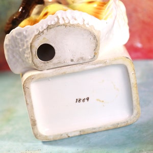 May include: White porcelain figurine of a woman in a yellow dress with a brown hat. The figurine has a hole in the bottom and the number 1869 is printed on the base.