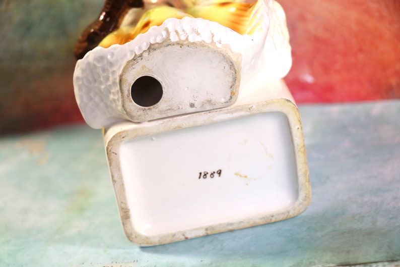 May include: White porcelain figurine of a woman in a yellow dress with a brown hat. The figurine has a hole in the bottom and the number 1869 is printed on the base.