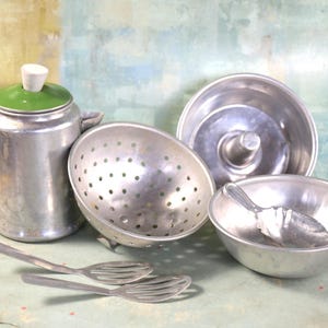 May include: A collection of vintage aluminum kitchenware, including a coffee pot with a green lid, a colander, a bundt pan, a bowl with a serving spoon, and two slotted spatulas. The items are arranged on a floral patterned surface.