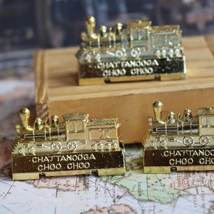 Op de afbeelding: Drie gouden speelgoedtreinlocomotieven met de woorden "Chattanooga Choo Choo" op de onderkant van elk. De treinen staan op een houten oppervlak en een kaart.