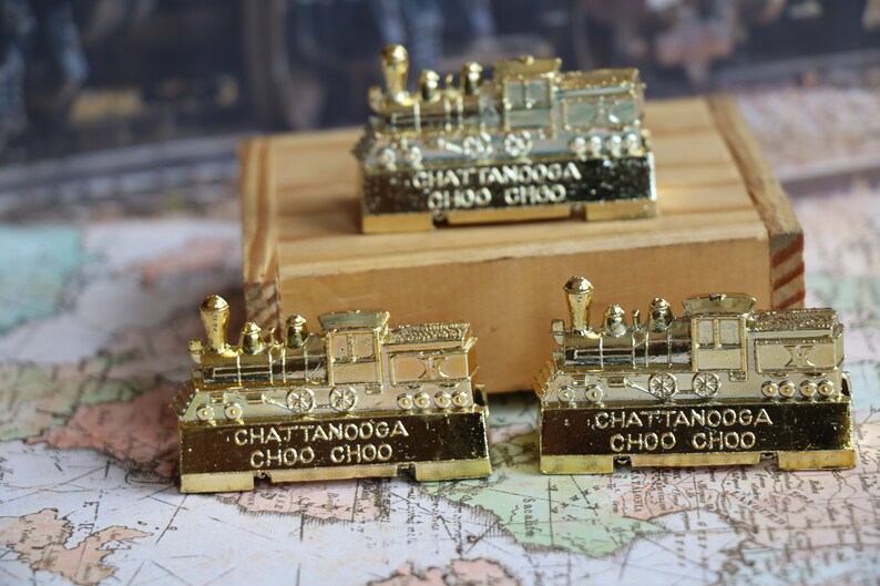 Op de afbeelding: Drie gouden speelgoedtreinlocomotieven met de woorden "Chattanooga Choo Choo" op de onderkant van elk. De treinen staan op een houten oppervlak en een kaart.