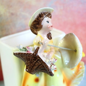 May include: A ceramic planter shaped like a girl holding an umbrella. The girl is wearing a yellow dress with white polka dots and a white hat. The umbrella is brown and white.