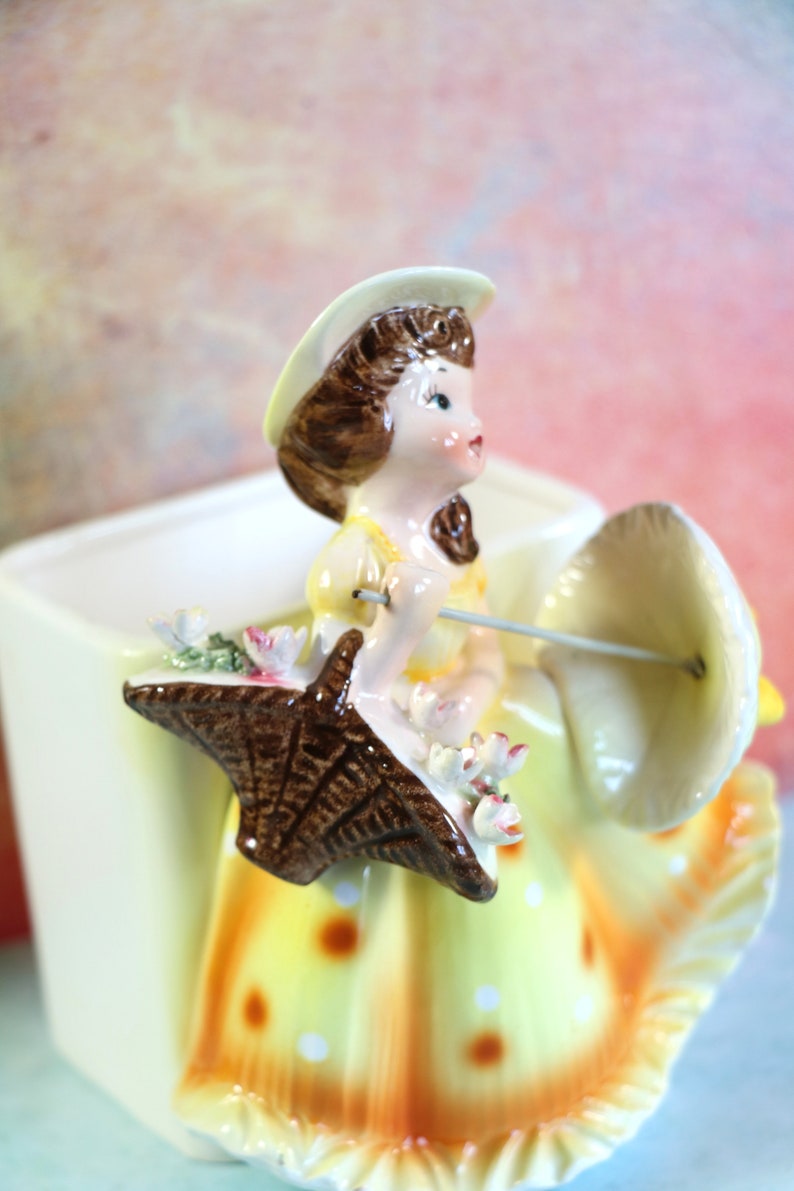 May include: A ceramic planter shaped like a girl holding an umbrella. The girl is wearing a yellow dress with white polka dots and a white hat. The umbrella is brown and white.